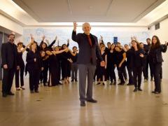 Foto dos integrantes do Coro de Câmara Comunicantus. Eles estão em uma sala. À frente do grupo, está regente e professor do coro. Ele é um homem mais velho, com cabelos curtos e brancos nas laterais da cabeça, usa um terno preto com gravata vermelha e está com uma das mãos levantadas. Ao fundo, está o grupo. Todos usam roupas pretas e olham para a câmera, com os braços levantados.