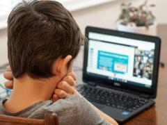Foto de um menino sentado assistindo à aula online. Ele tem a pele branca, cabelos curtos e escuros, usa uma camiseta cinza e está com o rosto apoiado nas mãos semifechadas. O fundo é desfocado, mas é possível observar um computador sobre a mesa. O computador mostra uma apresentação de slides dividindo espaço com uma videochamada.