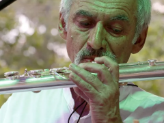 Foto de Toninho Carrasqueira, homem branco, idoso de cabelo brancos e bigode grisalho que usa uma camisa branca e toca uma flauta transversal. 