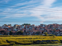 Foto de comunidade periférica próxima a um rio. Em primeiro plano, é possível observar parte do leito do rio e um campo de grama verde. Na parte superior, existem vários fios de alta tensão. Em segundo plano, há muitas casas próximas umas das outras, a maioria com tijolos e cimento aparentes. Existem poucas árvores no local. Ao fundo, um céu azul com algumas nuvens dispersas.