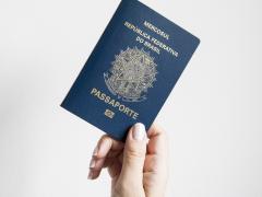 Foto. A frente de um fundo branco, uma mão, de uma pessoa branca, segura um passaporte, um livreto azul escuro com informações em dourado.