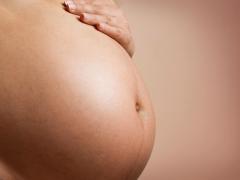 Foto de uma grávida. Apenas sua barriga, grande por conta da gravidez, aparece na imagem. A mão da mulher, que é branca, está apoiada em cima da barriga. O fundo da imagem é totalmente rosa.