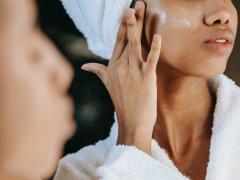 Foto de uma mulher negra com uma toalha no cabelo passando creme em frente ao espelho. No canto esquerdo da imagem, aparece o seu rosto desfocado. Em segundo plano e em foco, a mulher passa o creme em sua bochecha. É possível ver apenas seu nariz e sua boca. Ela veste um roupão de banho branco. 