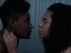 Foto de uma cena do curta. Duas mulheres negras se olham profundamente nos olhos. A da esquerda usa um brinco e tem cabelo crespo e curto. A da direita tem cabelo cacheado e volumoso. Ela coloca sua mão esquerda no pescoço da outra mulher. 
