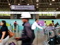 Foto do vai e vem de transeuntes com bagagens no terminal de passageiros do Galeão. Ao fundo, placa informativa  com o ícone de um avião subindo e os dizeres "Embarque doméstico" em português e inglês. 