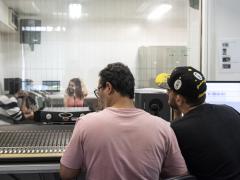 Foto de dois homens mexendo em uma mesa de mixagem de som. Eles estão de costas para a câmera. O da esquerda usa óculos e veste uma camiseta lilás, enquanto o da direita usa um boné e uma camiseta pretos. Ao fundo, por trás de uma tela de vidro, encontram-se duas mulheres e um homem gravando um material sonoro. À frente de cada um deles encontra-se um microfone.