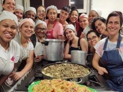 Foto de diversas mulheres ao redor de um fogão. Todas olham para a câmera e sorriem. A maioria usa toucas prendendo os cabelos. Em cima do fogão há grandes travessas com alimentos prontos e duas panelas.