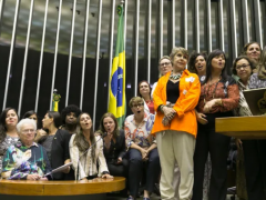 Foto de diversas mulheres, de diferentes idades e de maioria branca, na mesa e no púlpito da Câmara dos Deputados. Elas estão em pé, mas algumas estão mais altas do que outras devido ao desnível entre a mesa e o púlpito. Atrás das parlamentares está hasteada uma bandeira do Brasil.