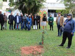 Foto de um grupo de pessoas em um gramado. Em primeiro plano, uma muda de árvore que acaba de ser plantada ao lado de uma placa em memória de Ciro Marcondes. Ao lado, um homem negro de barba grisalha e dreadlocks na altura do ombro fala. Ao fundo, homens e mulheres de diferentes idades observam a cena atentos. 