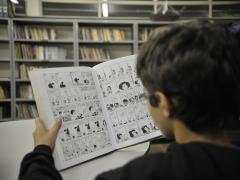 Foto de um menino branco, com cabelos curtos e castanhos que está sentado de costas. Ele lê um livro de histórias em quadrinhos. Ao fundo, estantes de livros.