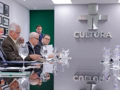 Foto do vice-presidente da República Hamilton Mourão sentado à mesa com diversos homens. Todos são brancos e têm cabelos grisalhos, à exceção de Mourão, que tem cabelos escuros e pele mais escura. Na parede ao fundo a logomarca da TV Cultura. 