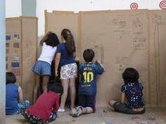 Foto de seis crianças desenhando e pintando em um mural de papel colado nas paredes e no chão. Algumas delas estão sentadas e outras estão em pé. 