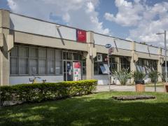 Foto da fachada do Departamento de Jornalismo e Editoração. Em primeiro plano, há um gramado ladeado por alguns arbustos. Mais ao fundo, está o prédio, onde encontram-se as placas do CJE e uma outra da ECA, que indica as direções para os outros departamentos. 