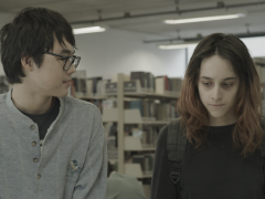 Imagem do filme Diário de Viagem. Um rapaz oriental de óculos observa menina de cabelo liso e pele pálida e com olhar baixo. Ao fundo uma prateleira de livros indicam que estão em uma biblioteca 