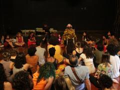Foto de várias pessoas sentadas em círculo, em cima de um palco de madeira. Elas se reúnem em volta do Mestre Martelo, que está sentado em uma cadeira. Ele é negro, usa calça e camisa estampadas nas cores amarela e preta, e um chapéu redondo e claro com uma faixa vermelha.