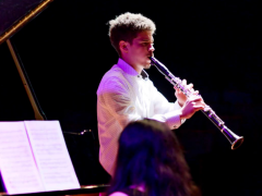 Foto de um homem tocando clarinete. Ele é branco, tem cabelos curtos e escuros e veste uma camisa branca. O fundo da imagem é totalmente preto, e o jovem é iluminado por luzes branca e roxa. Na porção inferior da foto, aparecem a cabeça de uma mulher que está de costas para a câmera e uma partitura apoiada no piano que ela toca.