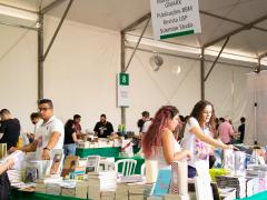 Foto do estande da editora Com-Arte na Festa do Livro da USP.  No expositor, os livros estão dispostos em uma mesa coberta por um tecido verde. Atrás dos livros, duas estudantes responsáveis pelo estade organizam as obras. Ao fundo, aparecem outros expositores do evento