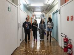 Foto de 4 estudantes caminhando juntos pelos corredores da ECA. Os 4 estão lado a lado. Da direita para a esquerda há um rapaz branco que tem cabelo curto e escuro. Ao seu lado há uma jovem de cabelo castanho claro, ondulado e pele branca. Em seguida há outra jovem de cabelo preto e pele branca. Mais à direita há uma jovem negra de cabelo cacheado. Ela segura uma garrafa plástica.