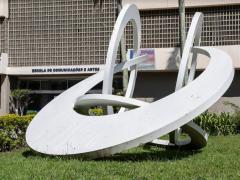 Foto de obra de arte sobre o gramado em frente à fachada do prédio principal da ECA. A escultura é branca, formada por 3 círculos e um semicírculo entrelaçados. Ao fundo, é possível ver a porta de entrada do prédio, com a inscrição Escola de Comunicações e Artes acima dela.