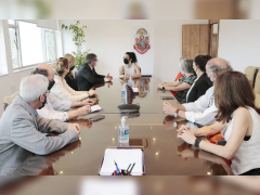 Foto de pessoas reunidas em uma grande mesa retangular. Na cabeceira da mesa, a cantora Marisa Monte - uma mulher branca, de cabelos escuros e ondulados, presos. Ela veste uma blusa branca e olha para a esquerda, onde está sentado o reitor da USP, um homem branco, de cabelos grisalhos e óculos. Ele veste um paletó cinza e uma camisa branca. Outras pessoas estão sentadas nos dois lados da mesa, homens e mulheres brancos. Ao fundo uma parede branca com o brasão da USP.