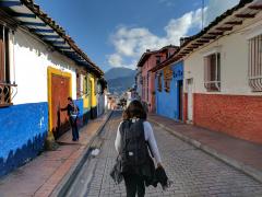Foto de uma jovem que caminha sozinha por uma rua de Bogotá, na Colômbia. Ela está de costas para a câmera e carrega uma mochila preta nas costas. Seu cabelo é castanho e liso. Ela usa uma blusa cinza, de manga comprida, uma calça jeans e tem um casaco amarrado na cintura. A rua por onde ela passa é feita de tijolos e tanto do lado direito, quanto do lado esquerdo, existem casas em estilo colonial, coloridas nas cores branco, azul, rosa, verde, vermelho e amarelo.