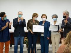 Foto de seis pessoas, três homens e três mulheres, em pé. Enquanto as mulheres seguram uma placa e um certificado, os homens aplaudem. Todos são brancos e usam máscara. Ao fundo, estão uma parede branca, uma mesa de madeira e uma bandeira azul hasteada. 