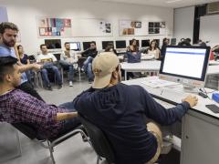 Foto de jovens do curso de Publicidade e Propaganda. São 20 estudantes sentados em uma sala, com cadeiras dispostas em círculo, entre homens e mulheres. Em primeiro plano, três jovens aparevem em frente a um computador, dois deles sentados e um de pé. A sala tem paredes brancas, com dois quadros com cartazes coloridos. Atrás dos estudantes sentados, em segundo plano, há dez computadores.