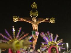  Foto de uma das alegorias apresentadas pela Estação Primeira de Mangueira durante o desfile das escolas de samba do Rio de Janeiro, no Carnaval de 2020. O carro alegórico faz uma releitura da história de Jesus Cristo, como um jovem negro baleado. Na imagem, o jovem está crucificado e olha para o céu. Acima, na cruz, lê-se a palavra "negro". Abaixo, aparecem membros da escola fantasiados, em primeiro plano mas desfocados. 