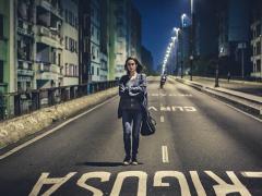 Cena do filme Segundo Tempo. Uma mulher jovem, branca de cabelos lisos e vestindo calça jeans, camiseta e blusa de manga comprida caminha de braços cruzados em uma das pistas do Elevado Costa e Silva, o Minhocão. À esquerda, fachadas de prédios quase coladas à borda da avenida. Ao fundo, uma fileira de postes iluminados e um céu azul escuro ao anoitecer. 