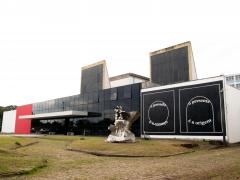 Foto da fachada do Espaço das Artes. Prédio térreo e retangular. à esquerda, uma pintura de formas geométricas, dois quadrados e circulos na cor branca em fundo preto, ao centro, o prédio possui vidro preto e na ponta, uma parede na cor vermelha. Uma obra de arte em metal está a frente do prédio.