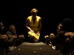 Foto de cena do espetáculo oresteia.br. Uma mulher está semi ajoelhada sobre uma mesa cercada por homens vestindo terno e usando máscaras. A mulher usa um vestido amarelo, tem pele clara e cabelos cacheados e curtos. A luz principal incide do teto, criando sombras e contrastes intensos.