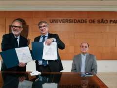 Foto do professor Paulo Nassar, um homem branco de cabelos e barba branca e óculos, e do reitor Carlos Gilberto Carlotti Jr, um homem branco de óculos e cabelos grisalhos, posando com exemplares do convênio entre ECA e Aberje. Ambos estão de pé atrás de uma mesa de madeira. Sentado na ponta da mesa, o professor Eugênio Bucci, um homem branco e calvo de cabelos escuros. Todos usam paletó e sorriem para a câmera.