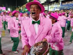 Foto de uma ala de sambistas de carnaval, com destaque para um homem negro, mais velho, tocando pandeiro e  cantando. Ele usa um terno rosa, gravata borboleta verde, camisa branca e chapéu rosa. O pandeiro tem uma imagem estampada. Os outros sambistas em volta cantam e usam a mesma roupa, incluindo uma calça verde e sapatos verde e rosa.