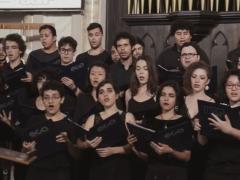 Foto de parte do Coral da ECA-USP, onde estão agrupadas várias fileiras de homens e mulheres cantando com uma pasta nas mãos