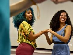 Fotos de duas mulheres segurando as mãos uma da outra. A mulher da esquerda é negra, tem cabelos crespos, compridos e azuis, veste uma blusa amarela e uma calça vermelha e usa brincos de argola. A mulher da direita também é negra, tem cabelos crespos, compridos e escuros, usa um vestido azul com um cinto preto e está sorrindo. Elas estão posicionadas uma de frente para a outra, mas olham para o lado. 