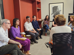 Foto de pessoas reunidas em um semi-circulo, parte delas sentada em um banco de madeira e encostados em um parede vermelha, outra parte em cadeiras de escritório. São três homens e quatro mulheres.