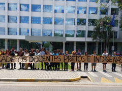  Foto de estudantes e professores em frente ao prédio da reitoria da USP. Vários estudantes, brancos e negros, estão alinhados lado a lado e seguram um cartaz bege em que está escrito "Cadê os docentes pretos e indígenas?”. Atrás dos estudantes, há um prédio espelhado