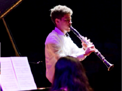 Foto de um homem tocando clarinete. Ele é branco, tem cabelos curtos e escuros e veste uma camisa branca. O fundo da imagem é totalmente preto, e o jovem é iluminado por luzes branca e roxa. Na porção inferior da foto, aparecem a cabeça de uma mulher que está de costas para a câmera e uma partitura apoiada no piano que ela toca.