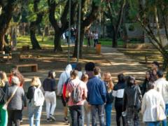  Foto tirada durante a visita monitorada à ECA. Na imagem, um grupo de estudantes passa pela parte externa da unidade, onde há várias árvores e bancos de concreto. Na foto, conta-se 19 pessoas andando de costas para a câmera.
