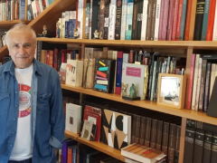 Foto de um homem posicionado em frente a uma estante de livros. Ele é branco, tem cabelos curtos e grisalhos e olhos escuros. Ele veste uma camiseta branca estampada e uma camisa jeans por cima. A estante é grande e ocupa toda a parede e, consequentemente, todo o fundo da imagem.