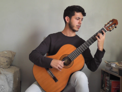 Foto de um rapaz tocando violão clássico e olhando para o instrumento. Ele é branco, tem cabelos curtos e escuros e veste uma blusa de manga longa preta e calça cinza. O jovem está em um quarto, sentado entre uma cama e uma escrivaninha. A parede ao fundo é cinza.