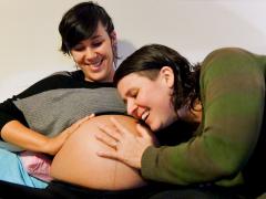 Foto de duas mulheres sentadas. A da esquerda é branca, tem cabelos curtos e escuros e veste uma blusa cinza e preta. Ela está grávida e sua barriga está exposta. A mulher da direita é branca, tem cabelos curtos e escuros, veste uma blusa verde e com o rosto próximo à barriga da companheira. As duas estão sorrindo e com uma das mãos na barriga grávida.