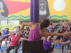 Foto de atividade no Sesc Pinheiros. Grupo de mulheres praticam atividade física. A professora está sentada em uma cadeira, de costas. Ela tem cabelos castanhos, ligeiramente ondulados, presos no topo da cabeça. Ela veste uma blusa roxa regata. à frente dela, está um grupo de mulheres sentadas em cadeiras, uma ao lado da outra. Elas repetem os movimentos da professora: um braço se estica para o lado, torcendo o tronco para o mesmo lado. o outro braço se mantém dobrado. Ao fundo, um painel artístico.
