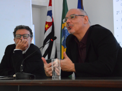 Foto de dois homens sentados em uma mesa. À esquerda, um homem de cabelos levemente grisalhos, usando óculos com haste escura, um terno preto e uma camisa branca, está com o queixo apoiado em uma das mãos. À direita, um homem calvo, de barba rala, usando óculos, um paletó preto e uma camisa escura fala ao microfone e gesticula com as mãos. Ao fundo, uma parede branca e as bandeiras do estado de São Paulo, do Brasil e da USP.