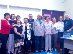 Foto de nove mulheres e três homens reunidos posando para uma foto de grupo. São todas pessoas idosas. Atrás das pessoas há uma porta dupla fechada, uma parede e é possível ver uma carteira de sala de aula. Também é possível ver uma mesa de professor com algumas vasilhas e sobras de comida. Aparentemente se trata de uma confraternização.