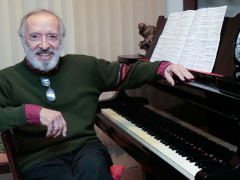 Foto de um homem branco, calvo, de cabelos e barba brancos. Ele usa um suéter verde escuro com punhos vermelhos e calça preta. Seus óculos de grau estão apoiados na gola do suéter. Ele sorri e posa ao lado de um piano de madeira escura, sobre o qual estende o braço esquerdo. Sobre a tampa do piano há uma partitura aberta..Ao fundo, uma persiana bege.