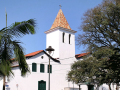 Foto da fachada de uma igreja em estilo colonial. As paredes são brancas, enquanto portas e janelas são da cor azul-marinho. Ao centro, a torre, que possui duas janelas estreitas e um telhado em forma de pirâmide com uma cruz no topo. A base da torre se liga, pelas laterais, a duas casas. Em primeiro plano, um poste de luz e as folhagens de uma árvore e de uma palmeira. Ao fundo, o céu azul, sem nuvens. 