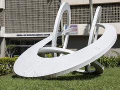 Foto de parte da fachada do prédio da ECA. Sobre um gramado verde, está uma escultura branca formada por quatro arcos que se entrelaçam entre si: um à frente, dois paralelos um ao outro e um quarto arco, mais próximo ao chão. Atrás da escultura, parte da fachado do prédio. A fachada tem elementos vazados, do tipo cobogó,  na cor bege. O nome da escola está afixado na parede, com as letras na cor azul escuro.