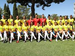 Foto oficial da Seleção Brasileira Feminina de 2019. 25 mulheres posam para uma foto, sendo 12 em pé e, à frente delas, 13 sentadas em um banco com as pernas cruzadas. Três das mulheres, que estão em pé e no centro da fotografia, usam um uniformes vermelhos, as demais usam uniformes amarelo e azul e meião branco. Elas estão em um gramado, a maioria sorri para a câmera e há um prédio branco de quatro andares no canto direito da foto. Árvores de diferentes formatos formam o segundo plano da imagem. 