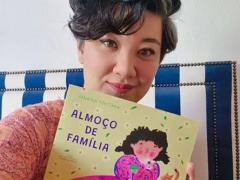 Foto da escritora Janaína Tokitaka, uma mulher de traços asiáticos, olhos castanhos, cabelos grisalhos, curtos e ondulados. Ela olha em direção à câmera e, com uma das mãos, segura seu livro Almoço de Família, cuja capa tem a ilustração de uma menina com cabelos cacheados e escuros, vestindo  blusa cor de rosa, em frente a uma mesa com pratos de comida. Ao lado da menina, na ilustração, há um gato.  Atrás de Janaína há um estofado com listras azuis e brancas e uma parede branca.